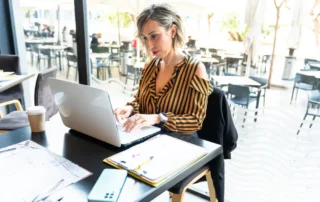 Femeie de afaceri care lucrează la laptop pe terasa unui restaurant, cu documente și notițe lângă ea, completând un brief pentru site web