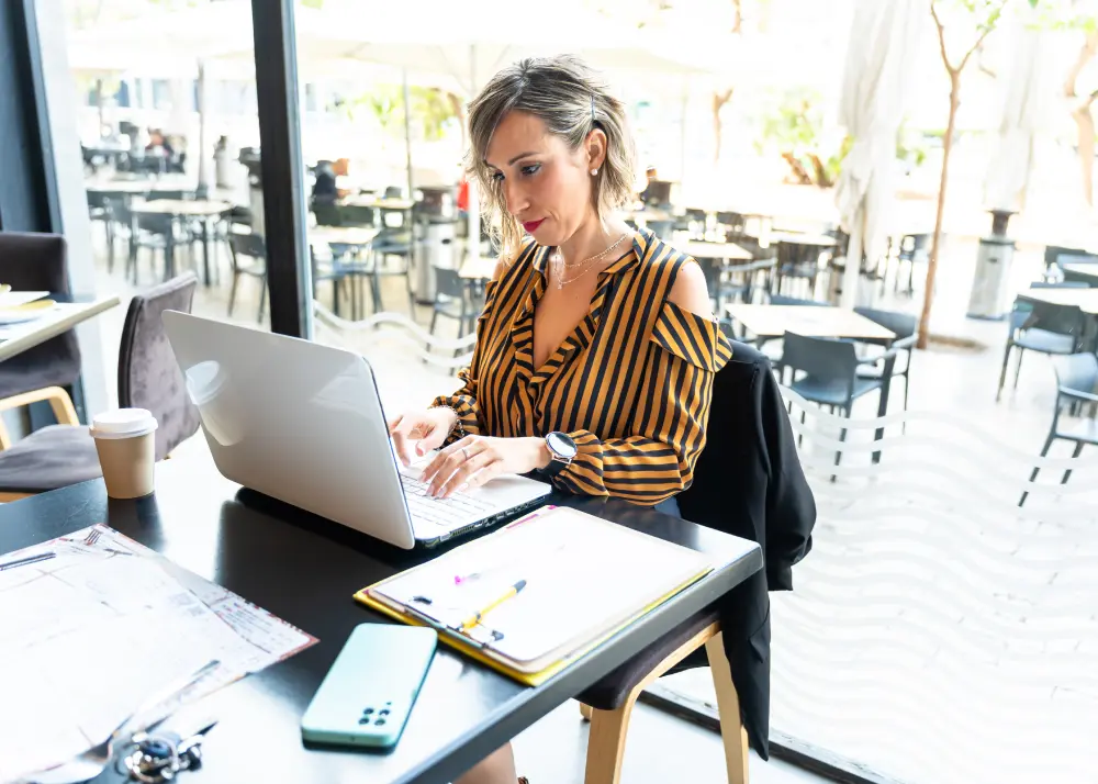 Femeie de afaceri care lucrează la laptop pe terasa unui restaurant, cu documente și notițe lângă ea, completând un brief pentru site web