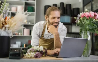 Florar zâmbind la laptop în magazinul de flori, gestionând comenzi și integrări pentru magazinul online pe WooCommerce