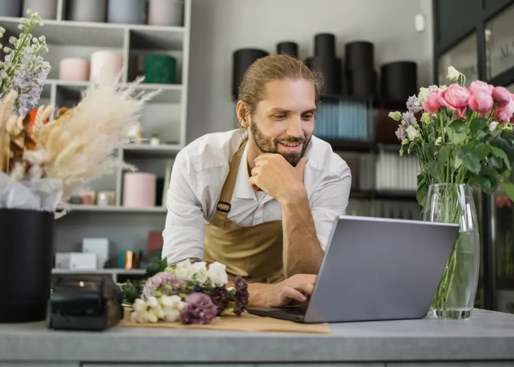 Florar zâmbind la laptop în magazinul de flori, gestionând comenzi și integrări pentru magazinul online pe WooCommerce