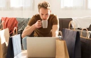 Bărbat care face cumpărături online de pe laptop, înconjurat de pungi de cumpărături
