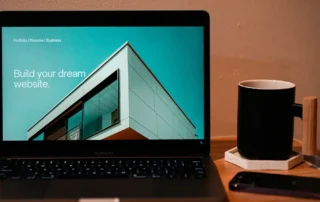 Laptop care afiseaza o platforma de creare site-uri cu mesajul "Build your dream website", langa o cana de cafea