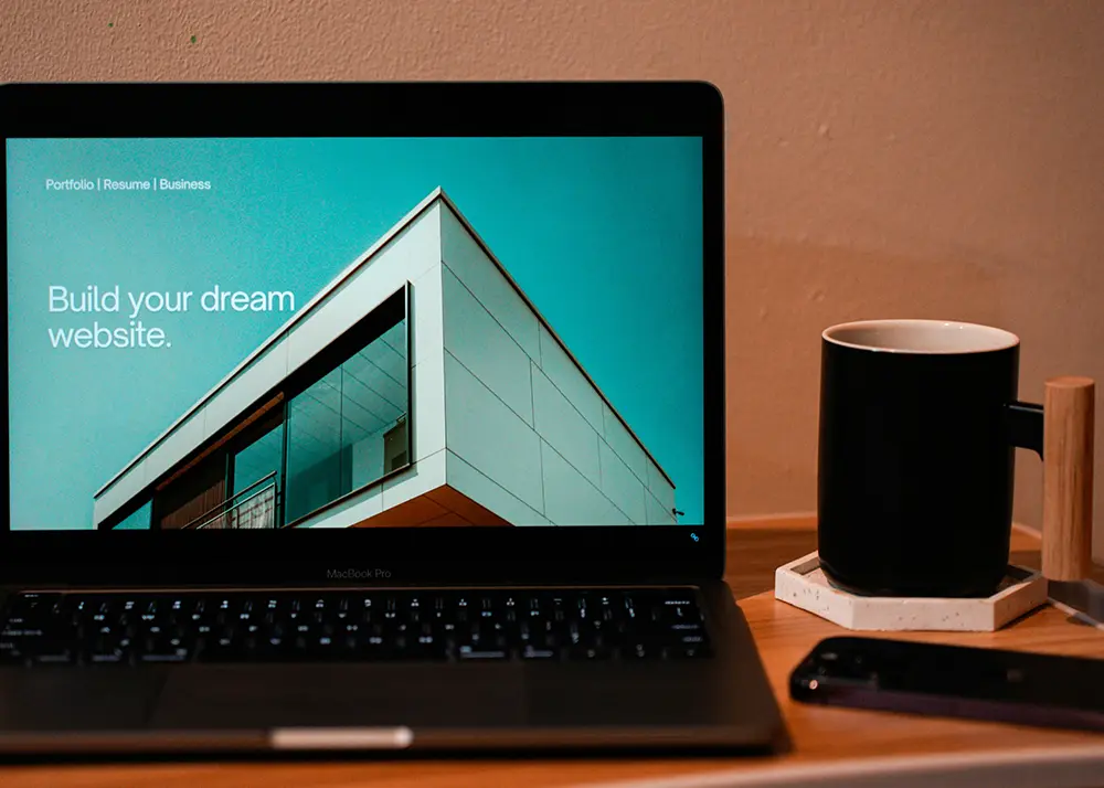 Un site se construieste usor, dar mentenanta lui necesita atentie continua Laptop care afiseaza o platforma de creare site-uri cu mesajul "Build your dream website", langa o cana de cafea