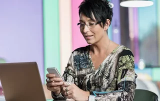 Antreprenoare care caută un furnizor pe telefon, în timp ce lucrează la laptop într-un birou modern