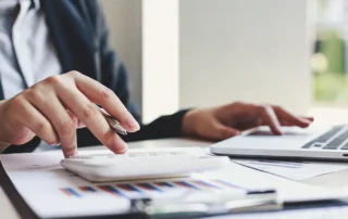 Antreprenor care calculează rentabilitatea investiției într-un site web, cu calculator, laptop și rapoarte cu grafice pe birou